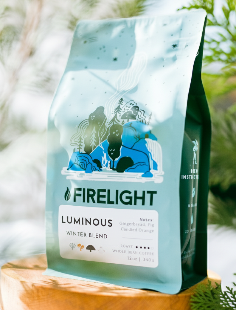 Bag of Firelight Coffee Luminous Winter Blend displayed on a wooden stand among green foliage, with soft natural light highlighting the blue packaging.