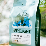 Bag of Firelight Coffee Luminous Winter Blend displayed on a wooden stand among green foliage, with soft natural light highlighting the blue packaging.