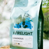 Bag of Firelight Coffee Luminous Winter Blend displayed on a wooden stand among green foliage, with soft natural light highlighting the blue packaging.