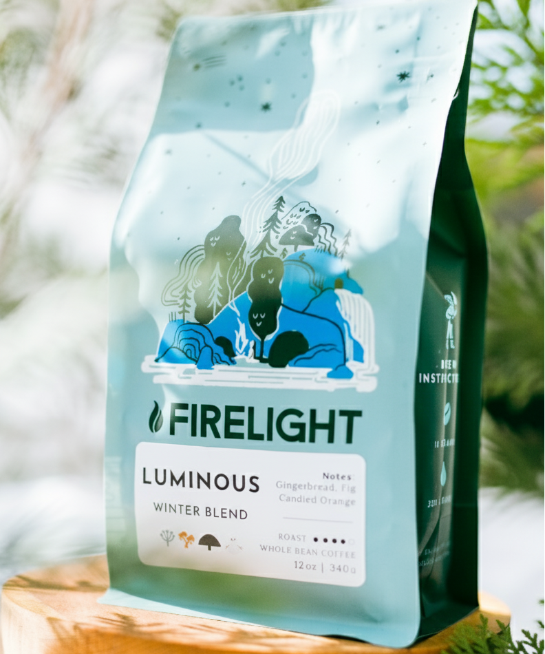 Bag of Firelight Coffee Luminous Winter Blend displayed on a wooden stand among green foliage, with soft natural light highlighting the blue packaging.